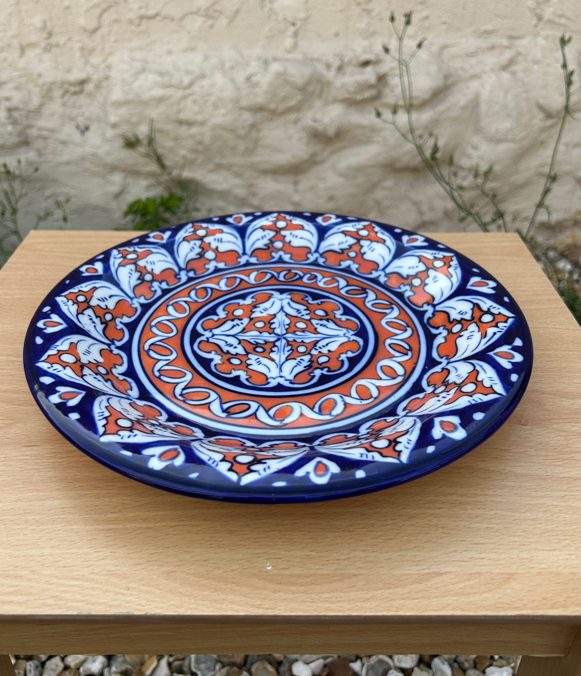 A round ceramic plate with a traditional Multani style hand-painted pattern in navy blue, terracotta orange, and white, displayed on a light wood table with a blurred stone wall background.